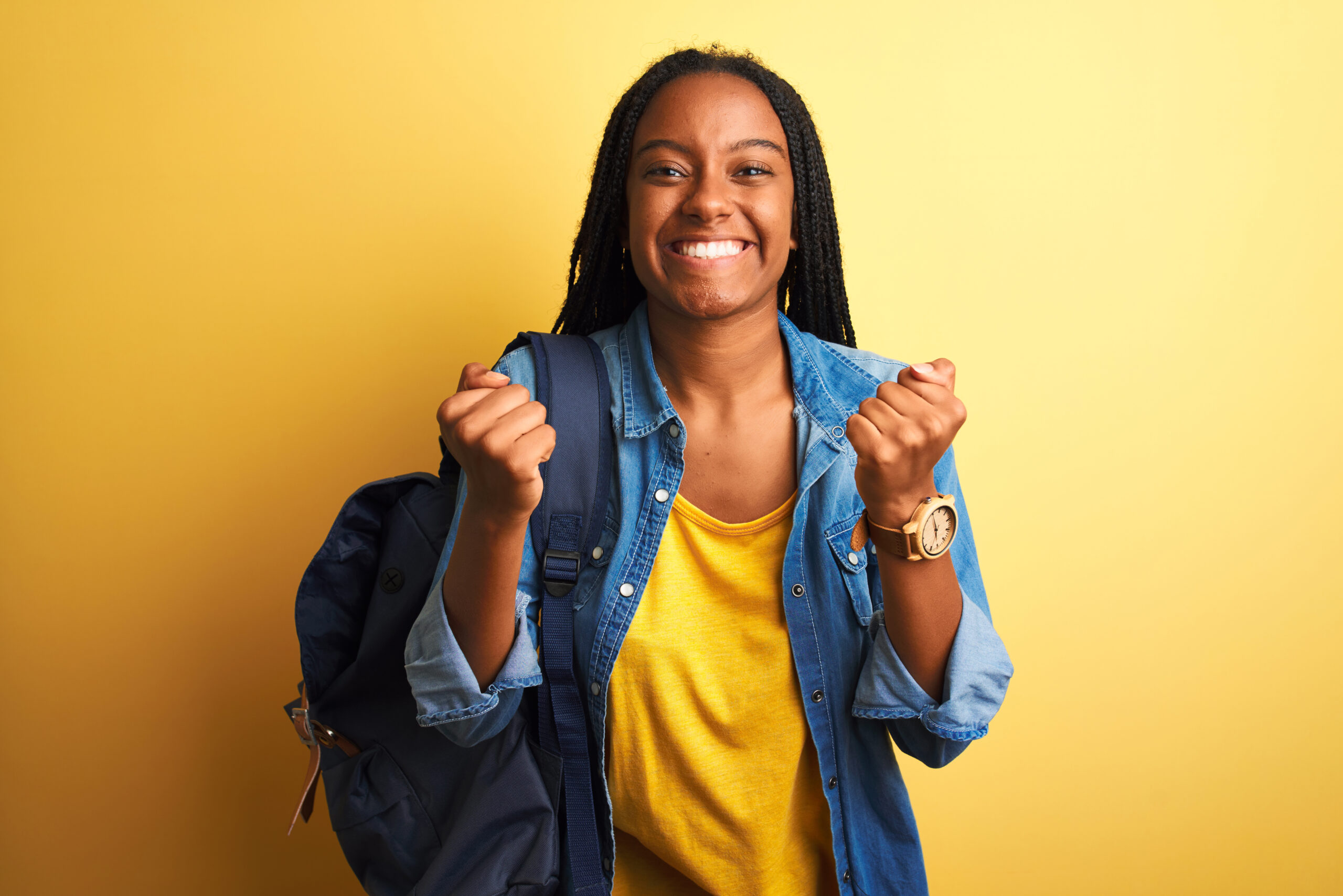 African american student woman wearing backpack standing over isolated yellow background celebrating surprised and amazed for success with arms raised and open eyes. Winner concept.